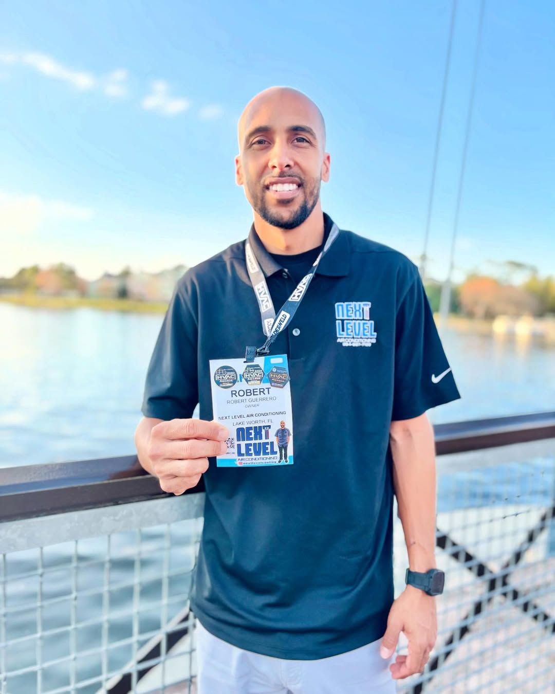 Owner in a branded black polo by a lake, holding up his AHR Expo attendee badge