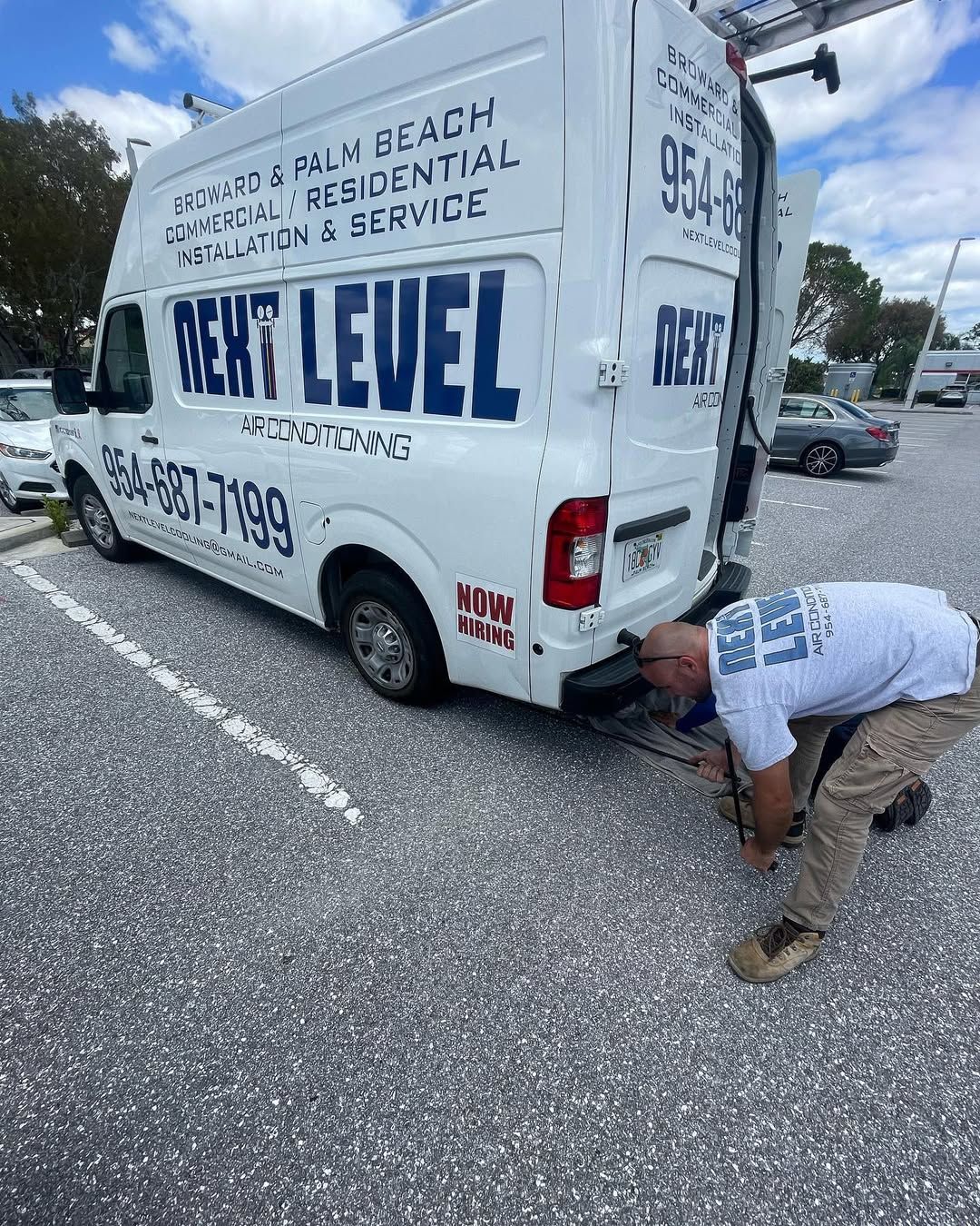 Branded van from the rear with a NOW HIRING decal, technician working at the open doors. Shows 954-687-7199