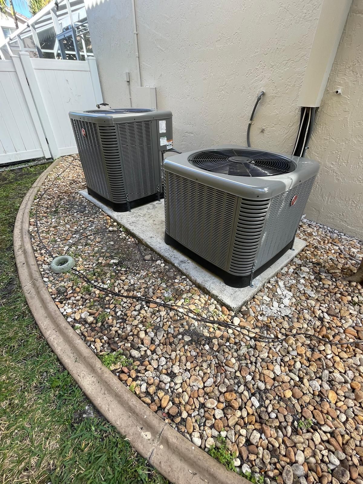 Two condensers on a shared pad against a white fence